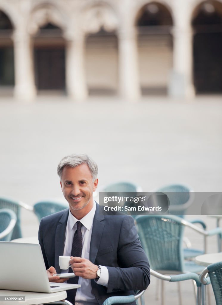 Portrait of smiling businessman drinking espresso and using laptop at sidewalk cafe