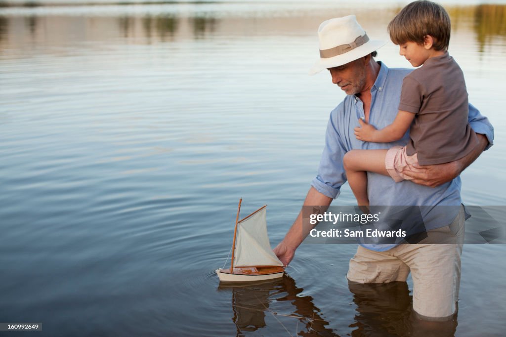 Grandfather and grandson wading in lake with toy sailboat