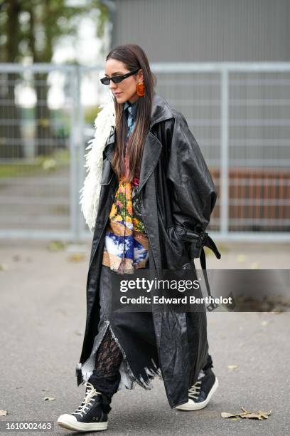 Guest wears black sunglasses, orange large earrings, a multicolored print pattern silk large shirt, a black shiny varnished leather long coat, a...