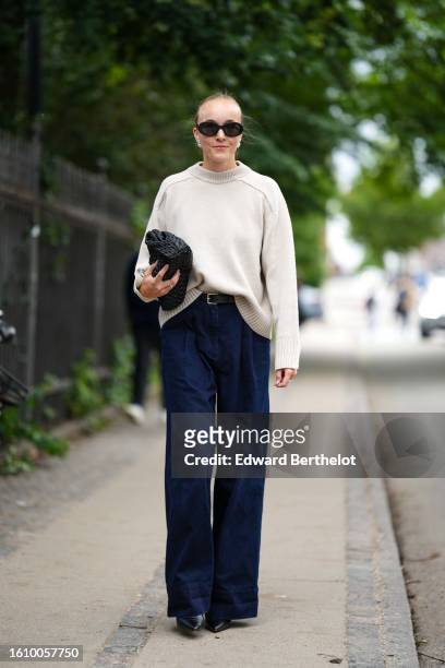 Guest wears black sunglasses, silver earrings, a beige wool pullover, a black braided shiny leather handbag, a black shiny leather belt, navy blue...