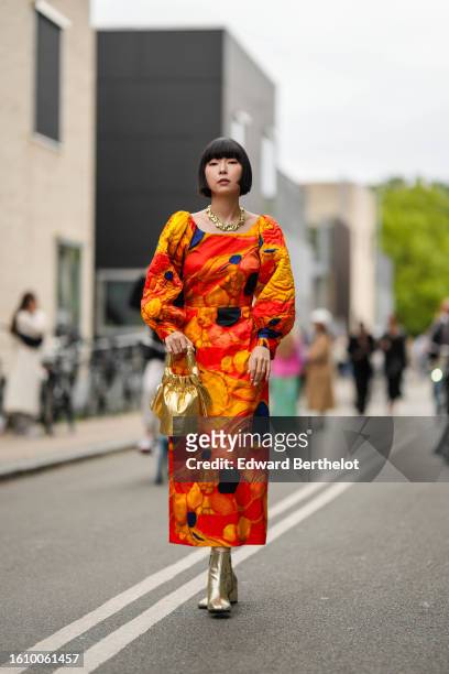 Guest wears a gold large necklace, a neon orange with yellow / navy blue print pattern long sleeves long midi dress, a gold shiny varnished leather...