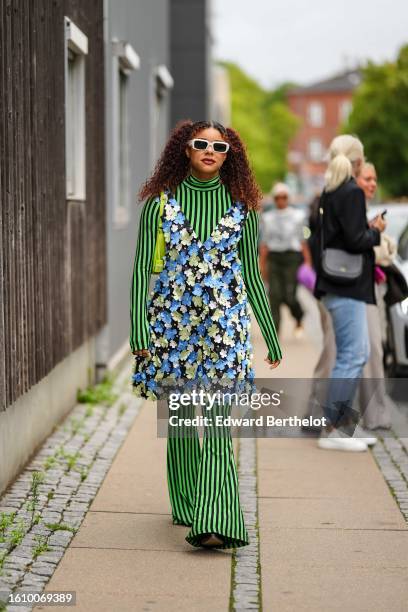 Guest wears white squared sunglasses, a neon green and black striped print pattern high neck / long sleeves / flared jumpsuit, a black / blue /...