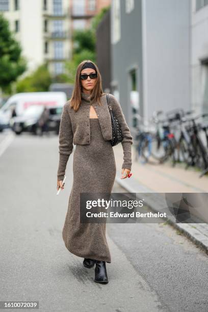 Guest wears a black large headband, black sunglasses, a dark brown felt wool cropped jacket, a matching dark brown felt wool shoulder-off long dress,...