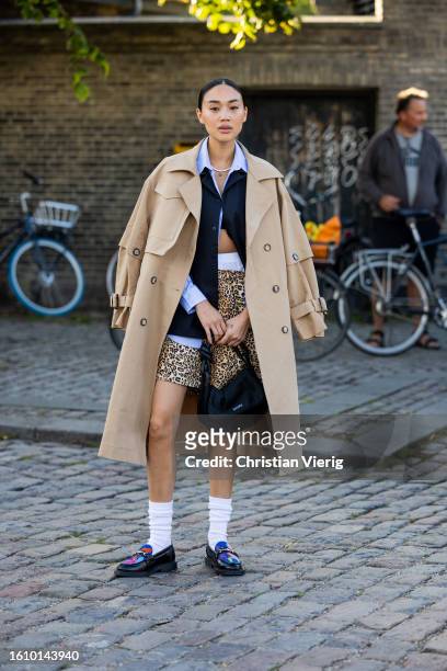 Guest wears animal print shorts, beige trench coat, blue button shirt, white socks, loafers outside Ganni during the Copenhagen Fashion Week...