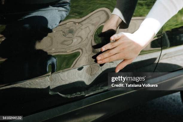 close-up of hand of lady touching dented car door, showing impact point and resulting creases and deformations in black metal. - scratched car stock pictures, royalty-free photos & images