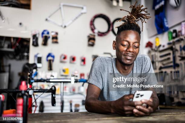 bike shop owner leaning on counter using his smartphone - generación del milenio fotografías e imágenes de stock