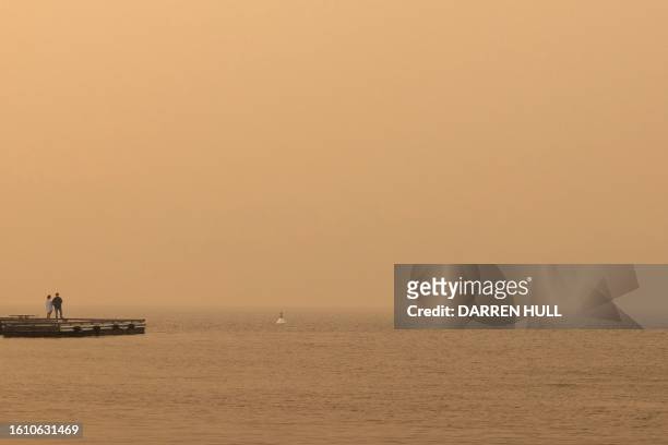 People stand on a dock as smoke fills the sky while wildfires continue to burn in Kelowna, British Columbia, on August 19, 2023. Around 30,000 people...