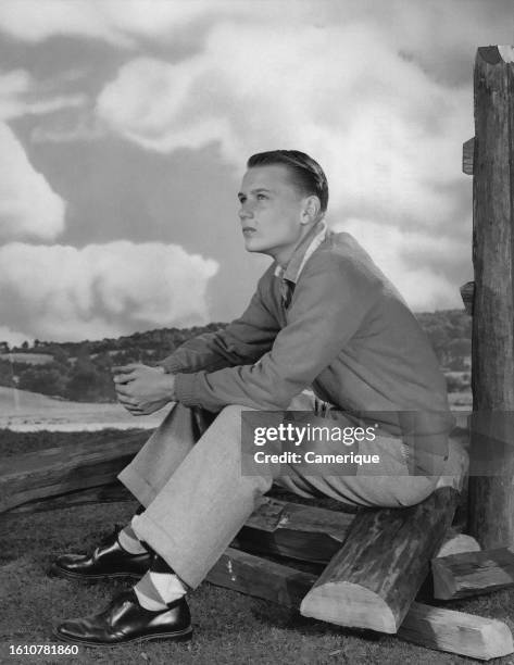 Young well-dressed man sitting on a pile of cut wood in a studio with his hands together lost in thought.