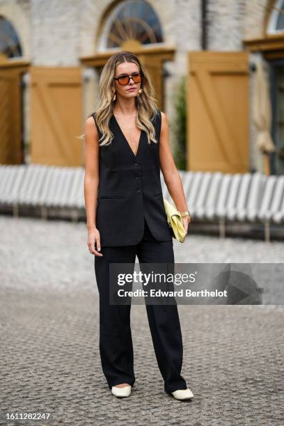 Guest wears black and orange sunglasses, gold large earrings, a black V-neck / buttoned sleeveless long gilet, black suit pants, a pale yellow matte...