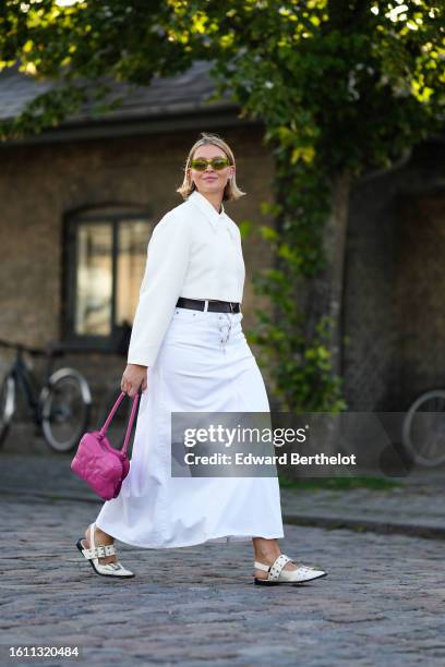 Guest wears green sunglasses, silver earrings, a white shirt, a black shiny leather belt, a white denim long skirt from Ganni, a neon pink shiny...