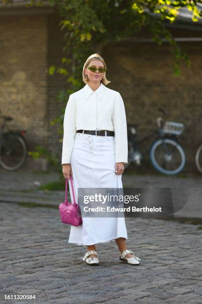 Guest wears green sunglasses, silver earrings, a white shirt, a black shiny leather belt, a white denim long skirt from Ganni, a neon pink shiny...