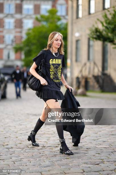 Guest wears silver earrings, a silver large chain necklace, a black with yellow print pattern t-shirt, a black pleated / accordion short skirt, black...
