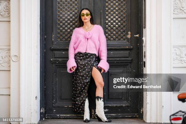 Guest wears green sunglasses, gold earrings, a silver large chain with beige teddy bear pendant necklace, a pale pink wool cardigan pullover, a black...