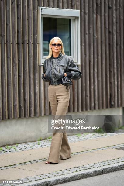 Guest wears black sunglasses, a black faded leather bomber coat, a black shiny leather shoulder bag from Chanel, brown high waist / flared pants,...