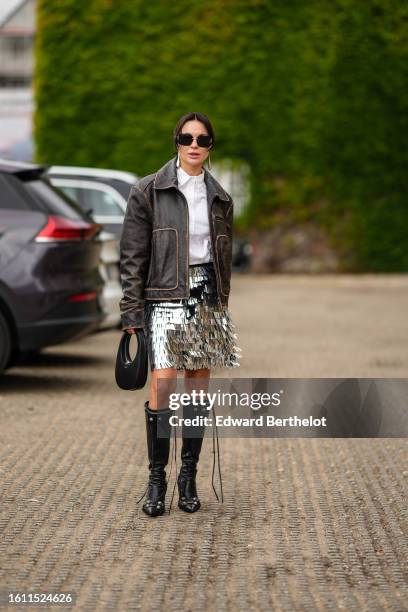 Guest wears black sunglasses, a white shirt, a dark brown faded leather zipper jacket, a silver embroidered metallic fringed short skirt, a black...