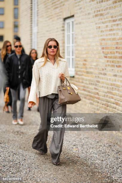 Guest wears gold earrings, a pale yellow braided wool polo shirt neck pullover, a brown grained leather Birkin handbag from Hermes, gray large suit...