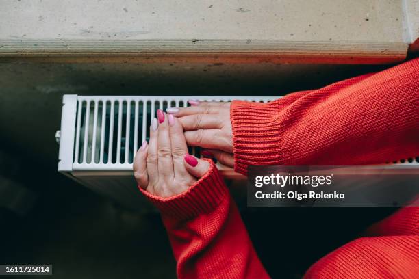 warmth in cold season for homeless people: cropped senior woman hands in red sweater leans on a heat radiator by the windowsill. top view - estufa eléctrica fotografías e imágenes de stock