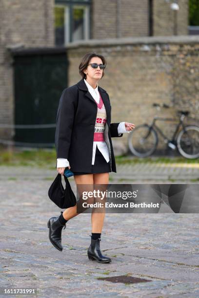 Guest wears black sunglasses, micro silver earrings, a white shirt, a red with white latte pattern wool V-neck pullover, a black blazer jacket, a...