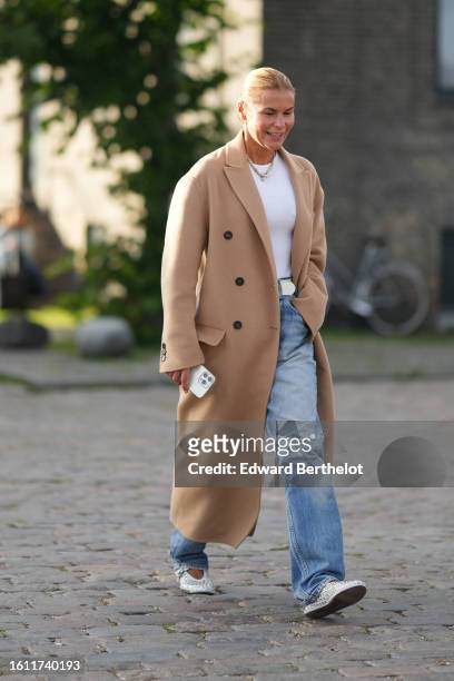 Guest wears a white t-shirt, a beige wool long coat, a white shiny leather belt, blue faded denim large pants, white leather nailed / studded...