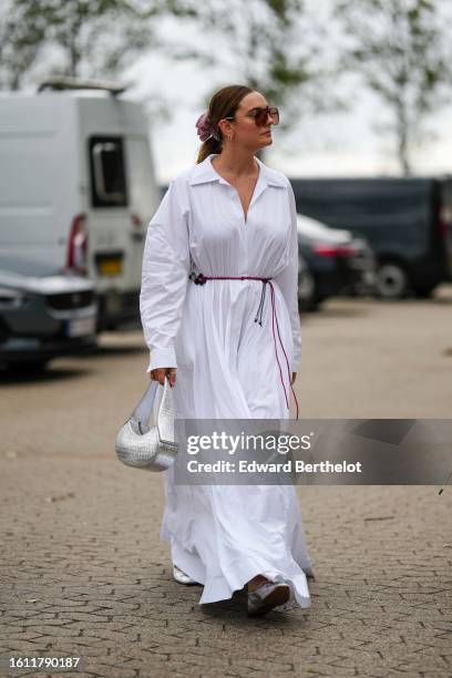 Guest wears brown large aviator sunglasses, gold earrings, a pink silk large hair elastic, a white shirt neck / long shirt dress, a silver shiny...