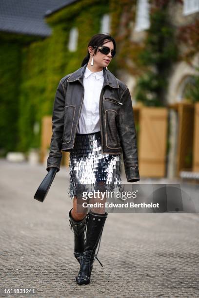 Guest wears black sunglasses, a white shirt, a dark brown faded leather zipper jacket, a silver embroidered metallic fringed short skirt, a black...
