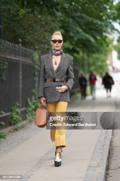 Guest wears black sunglasses, a black / beige / brown print pattern silk scarf, a gold chain with large pendant necklace, a gray wool blazer jacket,...