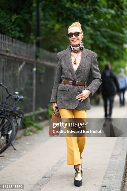 Guest wears black sunglasses, a black / beige / brown print pattern silk scarf, a gold chain with large pendant necklace, a gray wool blazer jacket,...