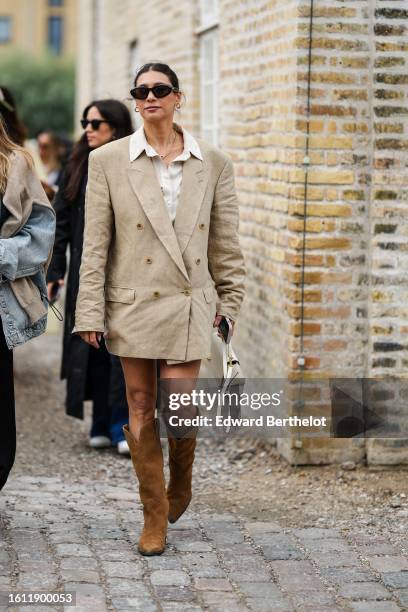Guest wears black sunglasses, a gold necklace, a white latte shirt, a beige oversized blazer jacket as a dress, camel suede pointed / block heels...