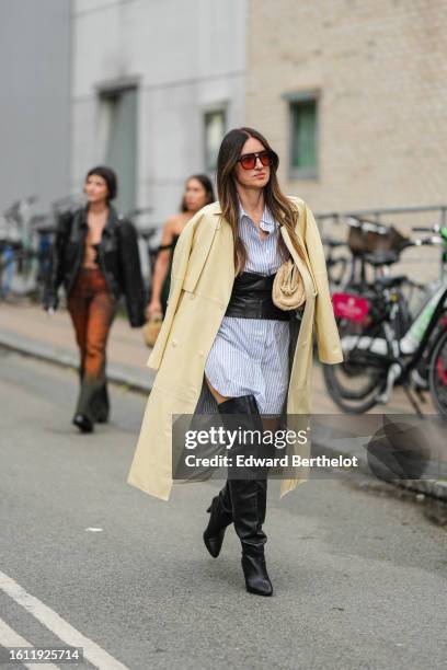 Guest wears red sunglasses, a blue and white striped print pattern oversized shirt dress, a black shiny leather large corset belt, a pale yellow long...