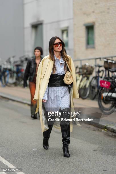 Guest wears red sunglasses, a blue and white striped print pattern oversized shirt dress, a black shiny leather large corset belt, a pale yellow long...