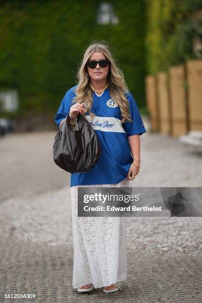 Guest wears black sunglasses, a gold pearls necklace, a blue with white print pattern sport t-shirt, a black shiny leather large handbag, gold...