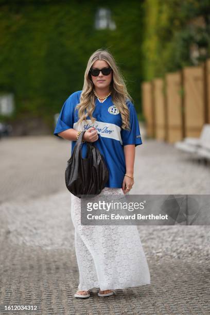 Guest wears black sunglasses, a gold pearls necklace, a blue with white print pattern sport t-shirt, a black shiny leather large handbag, gold...