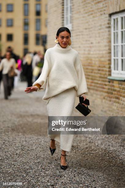 Guest wears gold large earrings, a white latte wool turtleneck pullover, a white latte wool long skirt, a black shiny leather handbag, black shiny...