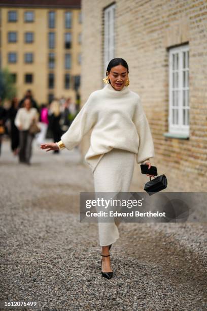 Guest wears gold large earrings, a white latte wool turtleneck pullover, a white latte wool long skirt, a black shiny leather handbag, black shiny...