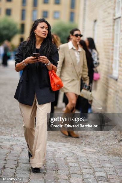 Guest wears gold earrings, a black t-shirt, a black blazer jacket, a neon orange satin handbag, a silver watch, silver bracelets, beige wide legs...