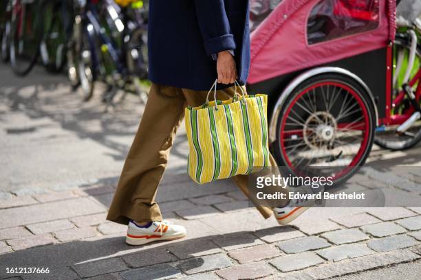 Guest wears a navy blue wool jacket, brown large suit pants, a yellow / white / green striped print pattern fabric handbag, white leather with orange...