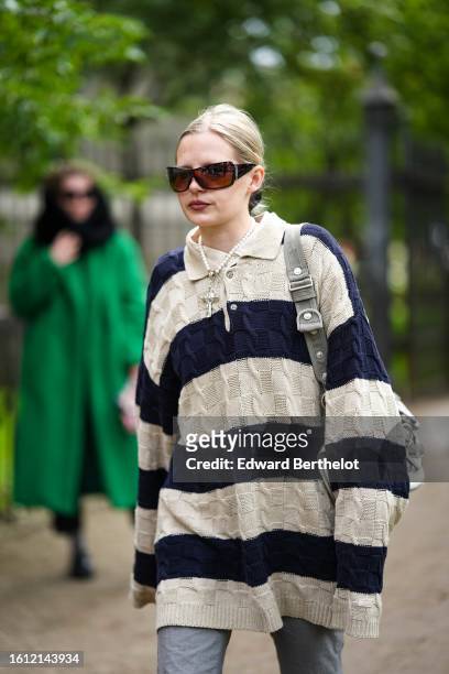 Guest wears black sunglasses, a beige shirt, a beige and navy blue braided wool striped print pattern oversized pullover, a gray shoulder bag, gray...
