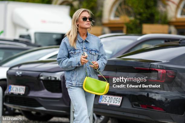 Guest wears black sunglasses, gold earrings, a pale blue faded denim oversized jacket, a green with yellow large puffy borders leather handbag, pale...