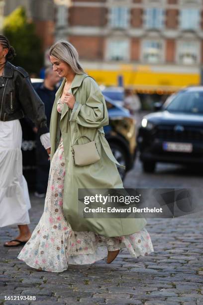 Guest wears a pale khaki long trench coat, a beige matte leather shoulder bag, a white latte with brown / pink flower pattern long dress, brown suede...