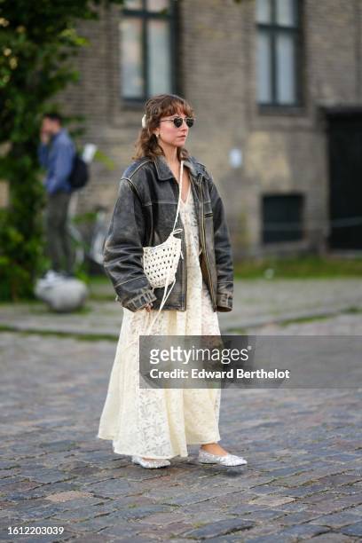 Guest wears black sunglasses, a black faded leather oversized jacket, a pale yellow lace print pattern long dress, a white braided wool crossbody...