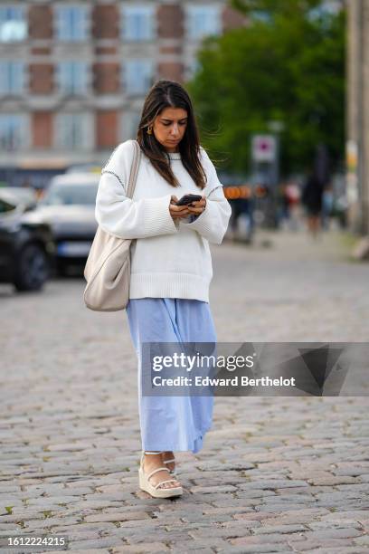 Guest wears gold earrings, a white wool oversized pullover, a beige matte leather large shoulder bag, a pale blue long dress, white leather strappy...