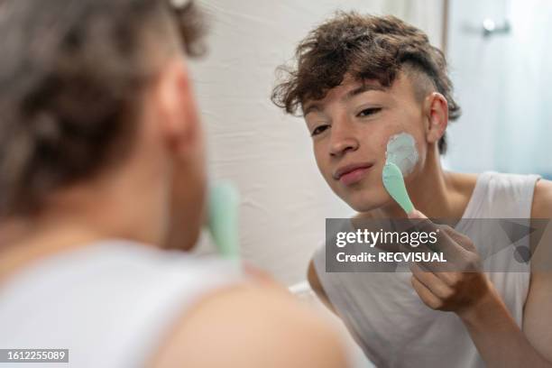 young man applying facial cream to his face and taking care of his skin in the bathroom. - skin care stock pictures, royalty-free photos & images