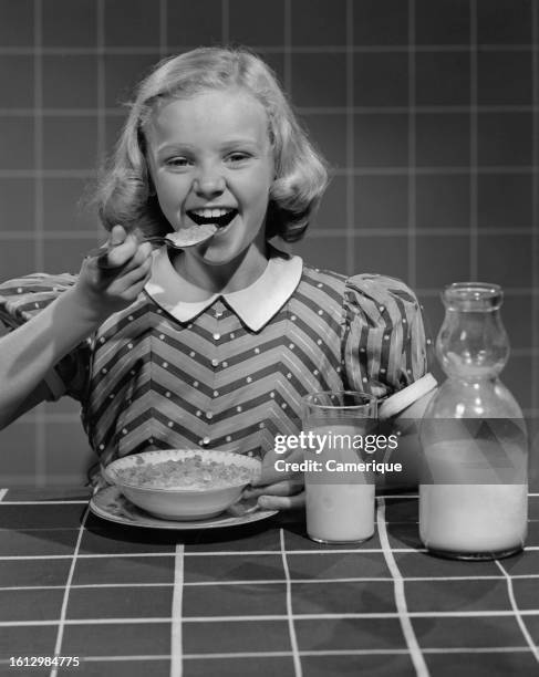 Girl in a dress sitting at the breakfast table putting a spoonful of cereal in her mouth as she gives a big smile.