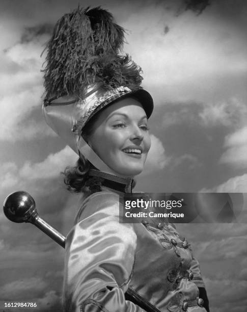 Headshot of a majorette against a cloudy backdrop wearing her shako cap which has a plume or pompom as it's decoration her baton under her arm.