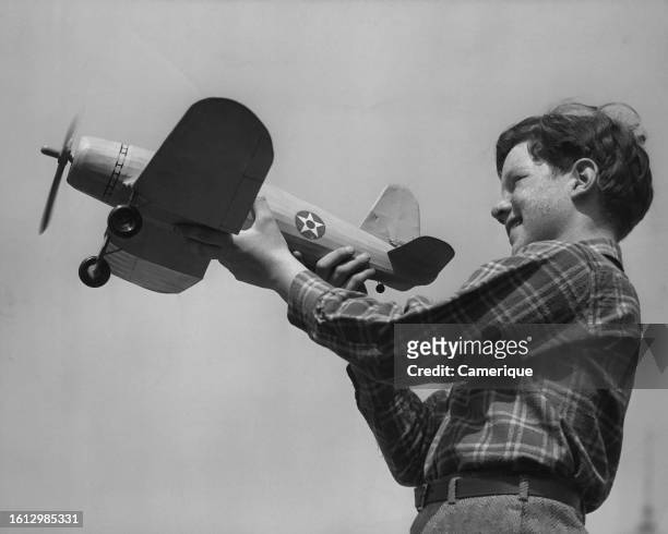 Freckle faced young boy wearing a plaid shirt holds a model airplane while the propeller is running prior to take-off.