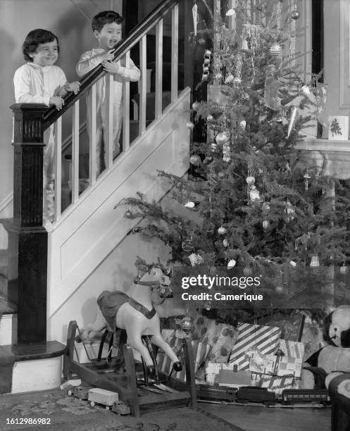 Young boy and his sister in pajamas standing on the staircase at the railing looking down on a Christmas tree loaded with toys and presents on...