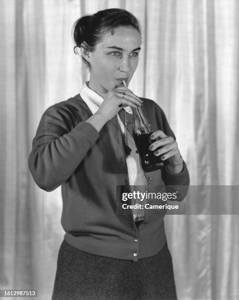 Teenage girl with her hair pulled back standing in front of a curtain drinking from a bottle of Coca-Cola with a straw.
