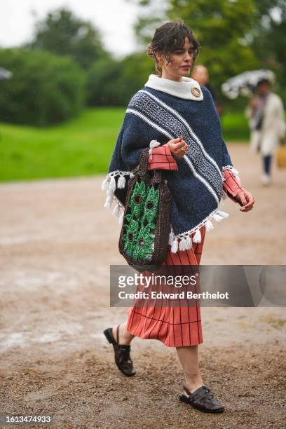 Guest wears gold large sun earrings, a navy blue braided wool with large white straps pattern and embroidered pompom borders cloak, a dark brown and...