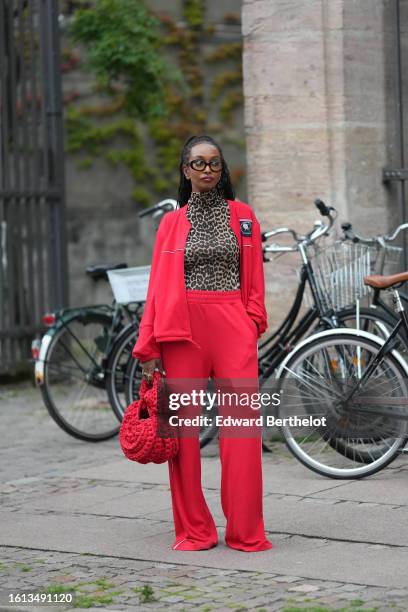 Guest wears black glasses, a brown / beige and black leopard print pattern turtleneck t-shirt, a neon red zipper sport sweater, matching neon red...