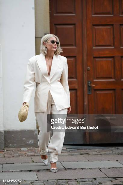 Guest wears black sunglasses, gold earrings, a white latte cut-out waist long blazer jacket, white latte suit pants, a beige braided leather Jodie...